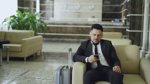 Pan Shot of Concentrated Businessman Using Smartphone Sitting on Armchair Inside Luxury Hotel with