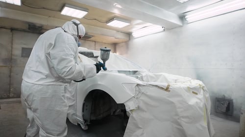 Big Man Is Wearing Protective Coverall with Hood and Respirator Is Painting Automobile Body After