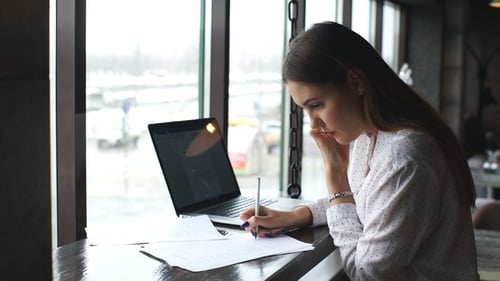 A Charming Young Student Is Looking at the Documents While Sitting in a Coffee-shop with a Laptop.