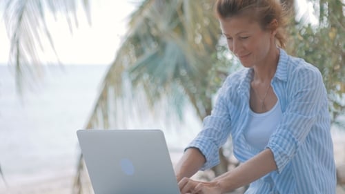 Young Woman Sitting at the Table with a Laptop with Sea View Behind