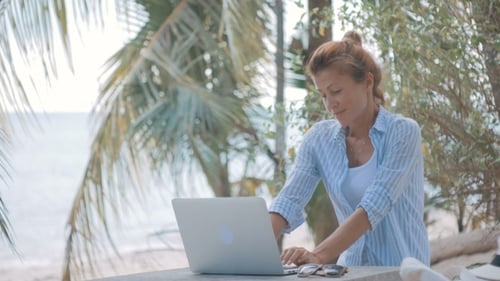Junge Frau sitzt am Tisch mit einem Laptop mit Blick auf das Meer dahinter, Kamera bewegen