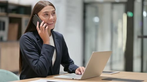 Businesswoman with Laptop Talking on Smartphone in Office