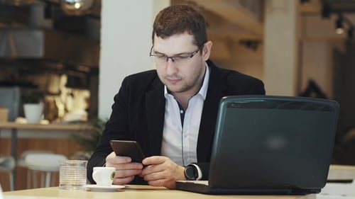 Man in a Business Suit Working on a Laptop and Mobile Phone in a Cafe