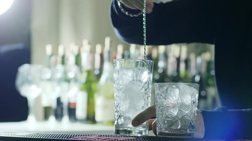 Bar Spoon in Hands of Barman Stir Ice in Glass on Bar Counter on Background of Bottles
