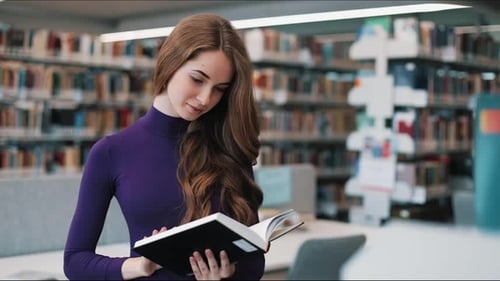 Thoughtful Young Female Student Reads a Book Standing in the Library