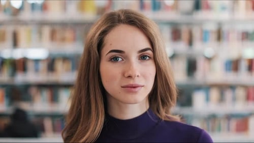 Smiling Female Student Holds a Book Standing Before the Shelves in the Library