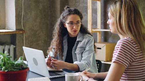 Young Enterpreneur Is Talking To Her Partner in Modern Office While Sitting at Desk Together. Women