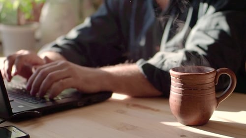 Teetasse und Laptop auf dem Tisch im Café. Die Tasse ist voll mit braunem Heißgetränk, das winkt.
EIN