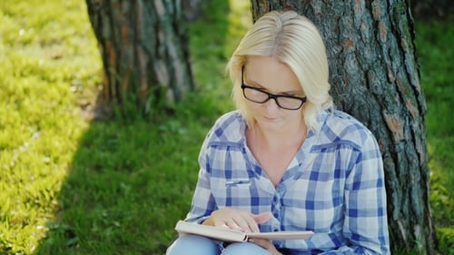 A Blonde Young Woman in Glasses Reads a Book in the Park. Sits Near a Tree, Beautiful Light Before