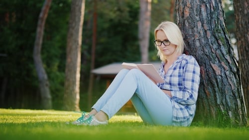 A Blonde Young Woman in Glasses Reads a Book in the Park. Sits Near a Tree, Beautiful Light Before