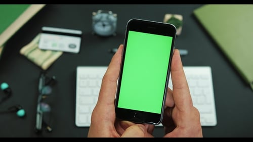 Man Holds Smartphone with Green Screen Over a Working Table