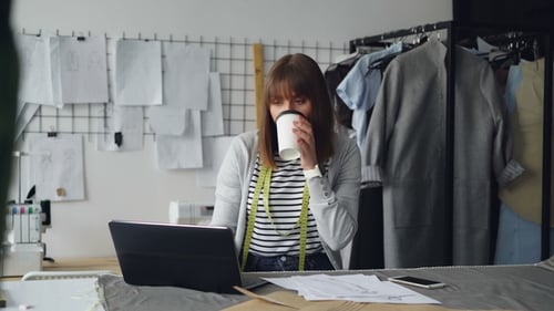 Junge attraktive Näherin arbeitet mit Laptop und trinkt Kaffee in ihrem Studio. Sie hat