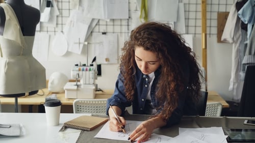 Young Female Artist Fashion Designer Is Drawing Women's Garment Sketch at Table in Modern Studio