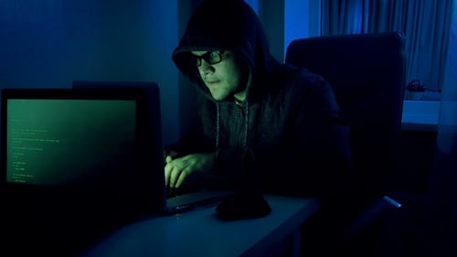 Young Male Pretends To Be Hacker. Geeky Man in Hood Working on Laptop at Night