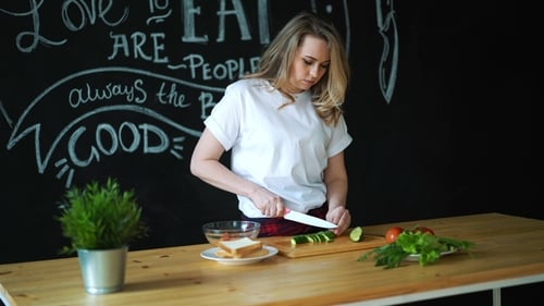 Beautiful Young Woman Cuts Green Cucumber with Kitchen Knife on Wooden Cutting Board