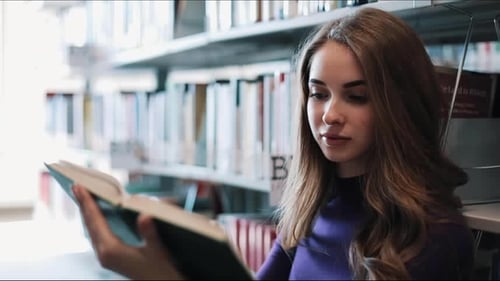 Thoughtful Young Female Student Reads a Book Standing in the Library