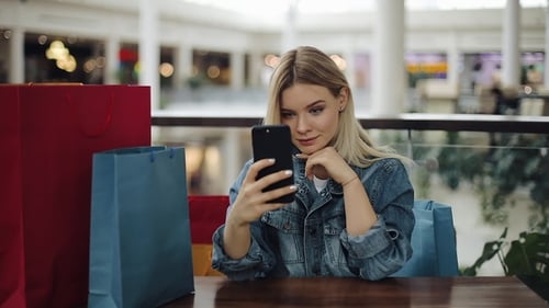 Pretty Blonde Girl Takes Selfie on Her Phone Sitting in Cafe with Shopping Bags