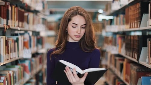 Thoughtful Young Female Student Reads a Book Standing in the Library