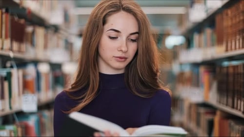 Thoughtful Young Female Student Reads a Book Standing in the Library