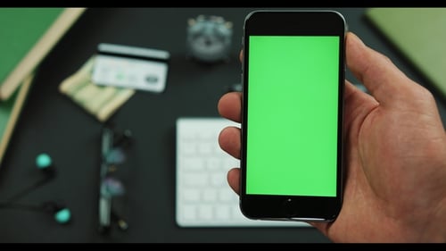 Man Holds a Black Smartphone with Green Screen Over a Working Table