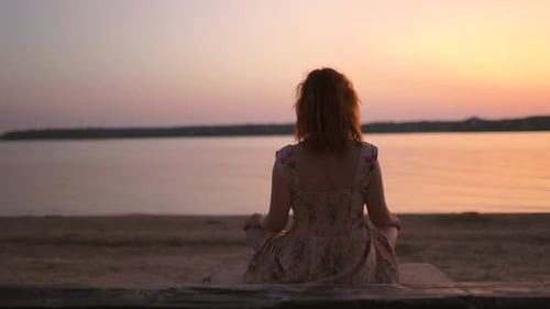 Young Woman Meditating at River During Sunset with Boat Passing By