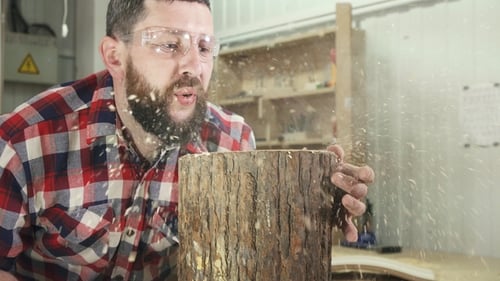 Man Carpenter in a Shirt with a Beard Blows Shavings in the Workshop