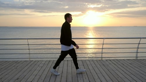 Silhouette of Young Trendy Modern Man Walking Along Wooden Embankment Near Sea or Ocean. Man in