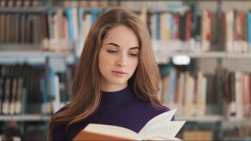 Thoughtful Young Female Student Reads a Book Standing in the Library