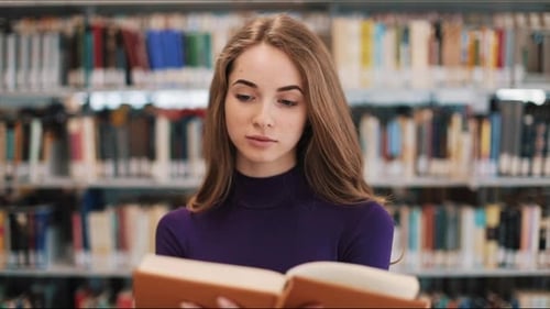 Thoughtful Young Female Student Reads a Book Standing in the Library