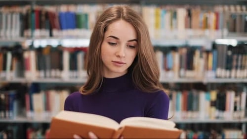 Thoughtful Young Female Student Reads a Book Standing in the Library