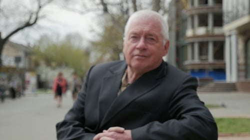 Wise Aged Man Sits on a Bench in a City Street in Spring in