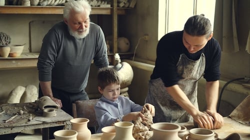 Professional Potter Is Kneading Clay on Worktable in Home Studio While His Son Is Helping Him
