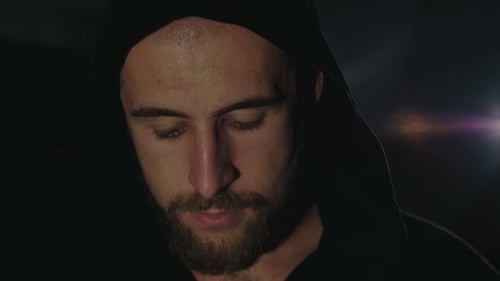 Strong Look at Camera of a Sweaty Man in Black Cloak in Darkness