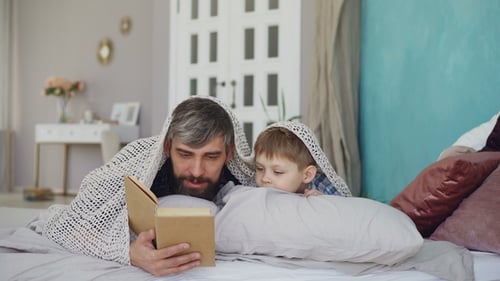 Father and Son Reading Under Blanket in Bed