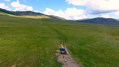 A View From the Air To Green Meadows, Hills in the Distance and a Car Riding Off-road