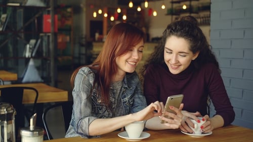 Two Young Pretty Girlfriends Are Using Smartphone in Cafe