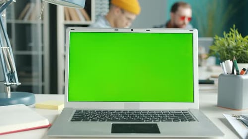 Chroma Key Green Screen Laptop on Desk with Employees Moving in Background in Office