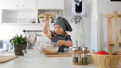 Preschool Girl Baker Kneads the Dough with a Large Wooden Spoon in a Bowl