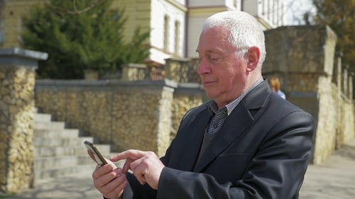 Retired Man Surfs the Net on His Phone in a Street in Spring in