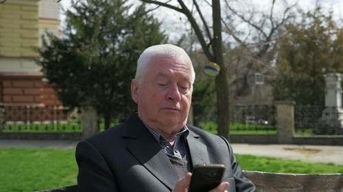 Senior Man Sits and Surfs the Net on His Smartphone in an Alley in Spring