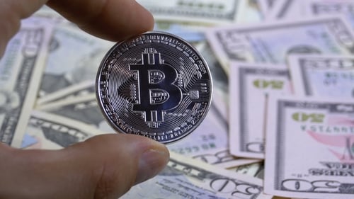 Hand in Finger hält eine Silbermünze Bitcoin, BTC auf einem Hintergrund mit Dollarscheinen