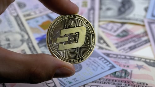 Hand in Fingers hält einen goldenen Münzstrich auf einem Hintergrund mit Dollarscheinen