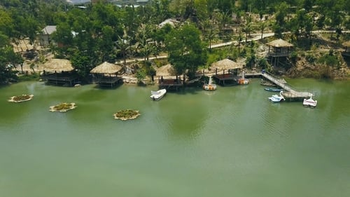 Wooden Bungalows with Thatched Roof and Boat Pier on Shore Green Lake in Resort Hotel Drone View