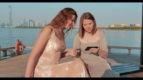 Two Smiling Girls in Light Dressesg Are Watching Photos on Smartphone. Blue Water Lake and Cityscape