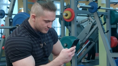 Young Man Using Phone While Having Exercise Break in the Gym