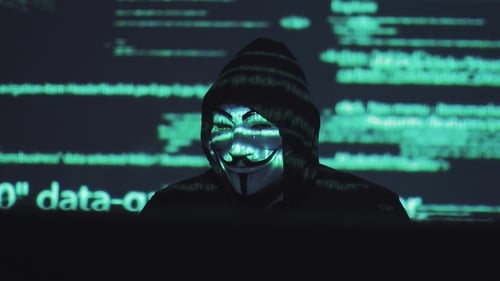 A Male Robber in a Mask Works on a Computer in a Dark Room. Computer Code Is Reflected on His Face
