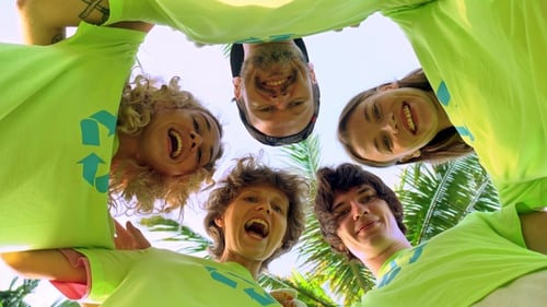 Five Young Volunteers in Green T-shirts with a Picture of Recycle Forming Huddles under Palm Trees