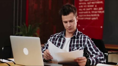 Happy Young Businessman Reading Paperwork at Desk in Office. Casual Man Smiling at Work. Man Working