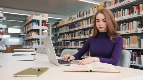Une étudiante travaille avec des livres et un ordinateur portable assise à la table de la bibliothèque