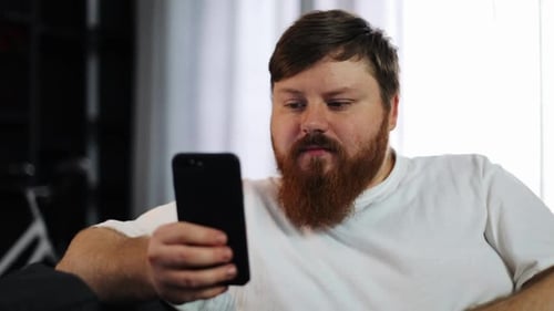 Serious Fat Man Works with His Smartphone Sitting on the Couch
